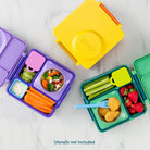 Three colorful ONIE bento lunch boxes arranged on a light marble surface, with food compartments filled with vegetables, crackers, and fruit. One box is open and one is closed, with a second box shown from above.