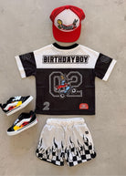 A children's birthday outfit set laid flat on a light concrete surface, including a black and white mesh jersey with 'BIRTHDAY BOY' and a large number 2 graphic, matching flame-print shorts, a red and white baseball cap, and black sneakers with yellow and white accents.