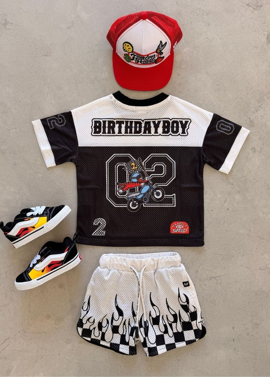 A children's birthday outfit set laid flat on a light concrete surface, including a black and white mesh jersey with 'BIRTHDAY BOY' and a large number 2 graphic, matching flame-print shorts, a red and white baseball cap, and black sneakers with yellow and white accents.
