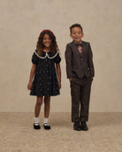 A young girl in a dark navy floral dress with a white Peter Pan collar and a red headband stands next to a young boy in a dark brown suit, bow tie, and dress shirt, posing against a textured beige studio backdrop.