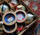 Decorative Christmas ornaments with wooden tops featuring Santa Claus and snowman designs, arranged in a box.
