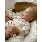 A baby lying on their back in a woven bassinet, wearing a cream-colored long-sleeve bodysuit with a pattern of orange foxes and small blue birds.