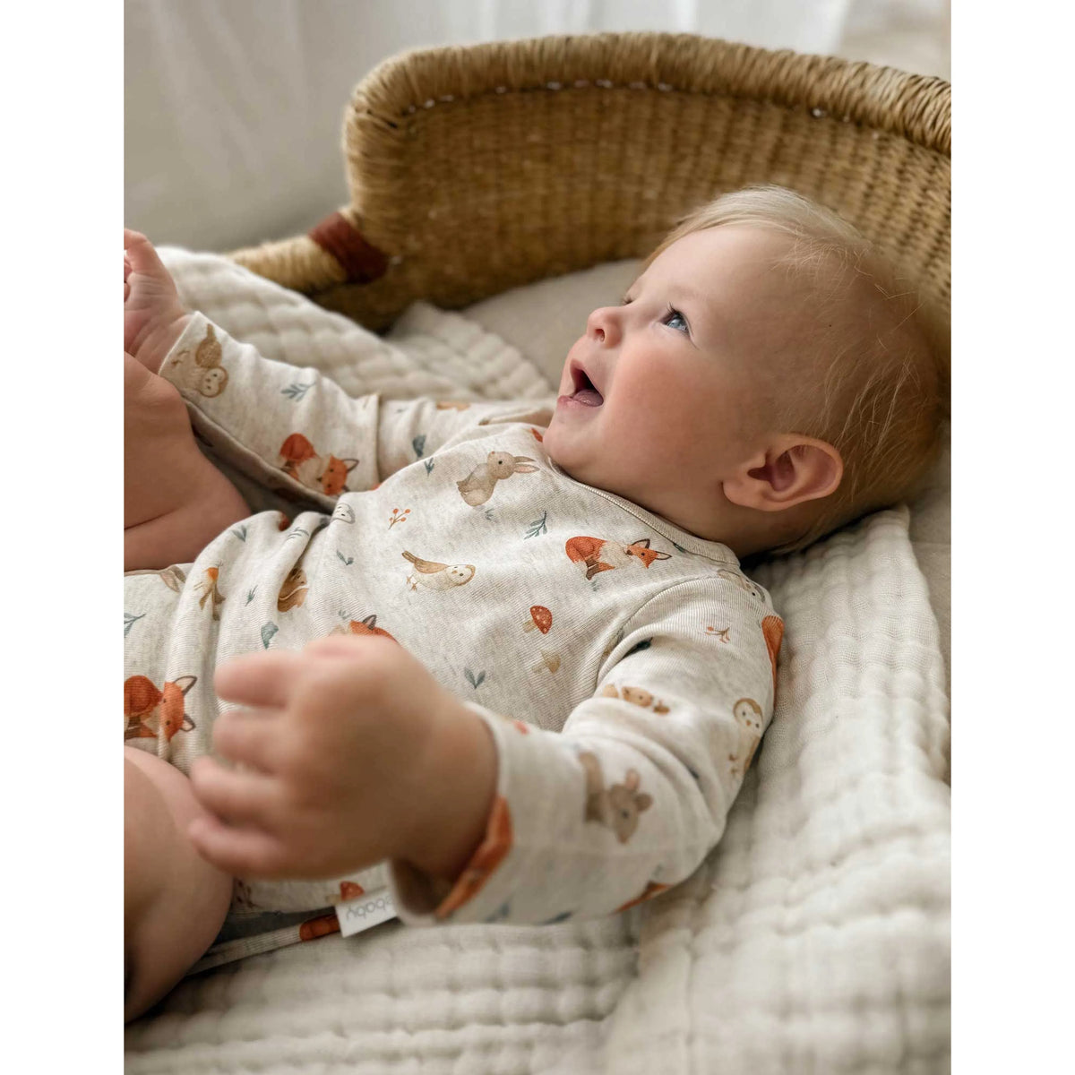 A baby lying on their back in a woven bassinet, wearing a cream-colored long-sleeve bodysuit with a pattern of orange foxes and small blue birds.