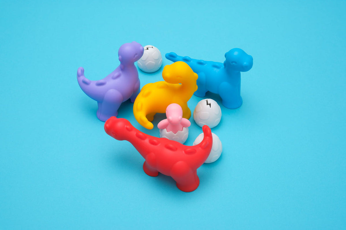 A set of colorful dinosaur-shaped bath toys in purple, blue, yellow, and red, accompanied by small white egg-shaped pieces, arranged on a solid light blue background.