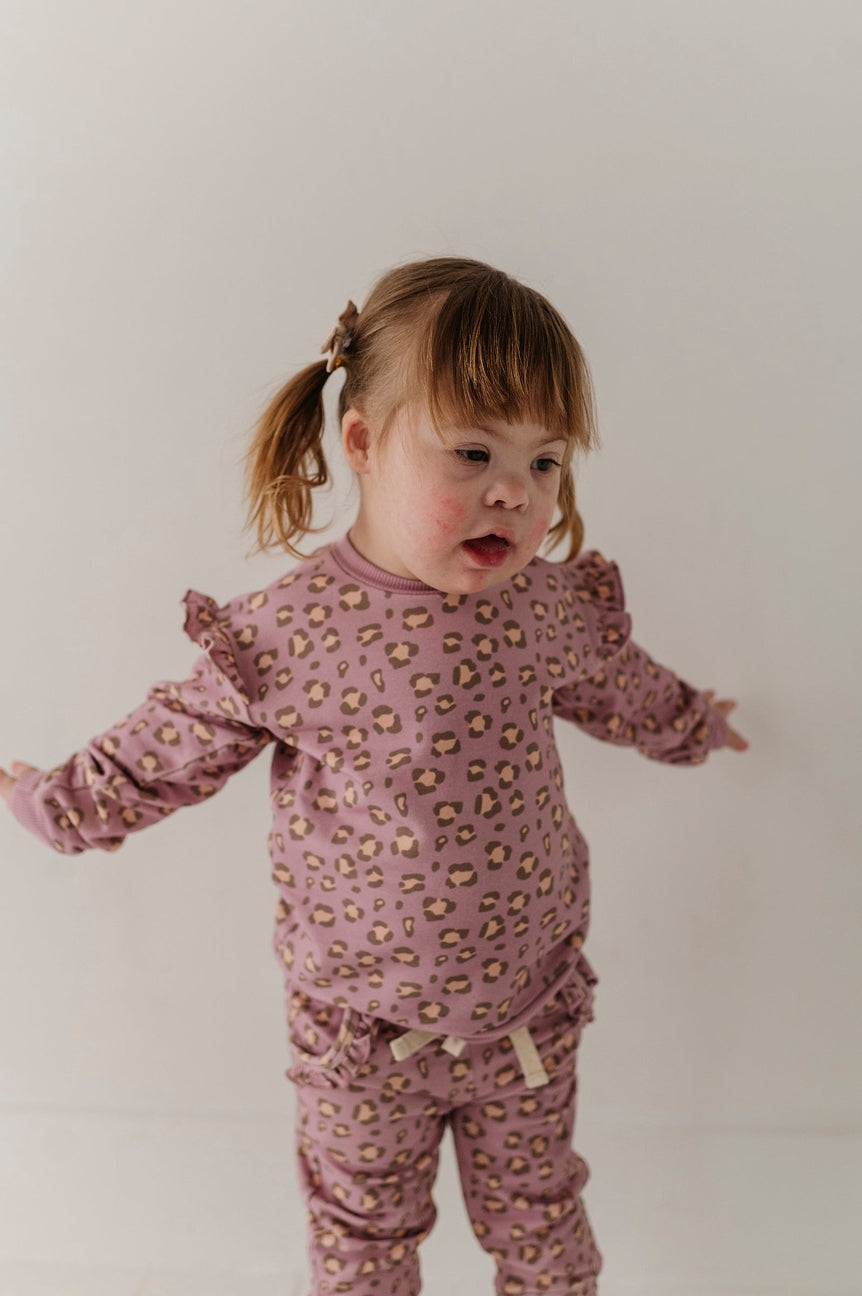 A toddler girl with brown hair in pigtails wearing a dusty pink long-sleeve pajama set with a small beige and brown animal print, standing against a plain off-white background.