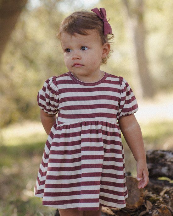 A toddler girl wearing a short-sleeve dress with horizontal burgundy and off-white stripes, standing outdoors in a blurred natural setting.