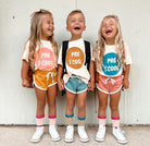 Three children standing together and laughing, wearing matching graphic t-shirts with colorful circular designs that read 'PRE S COOL' paired with different colored shorts, socks, and white sneakers against a plain light wall.