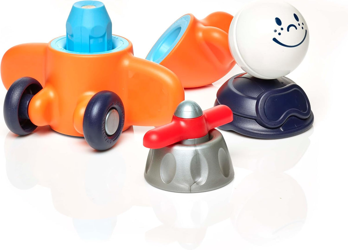 SmartMax My First Plane magnetic toy parts disassembled, including an orange airplane body, a blue connector piece, black wheels, a gray base, a red propeller, and a white ball figure with a smiling face on a white background.