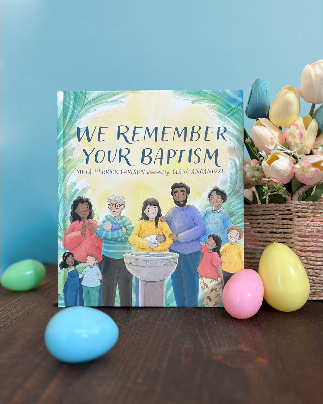 Book titled 'We Remember Your Baptism' with Easter eggs and a basket on a wooden surface.
