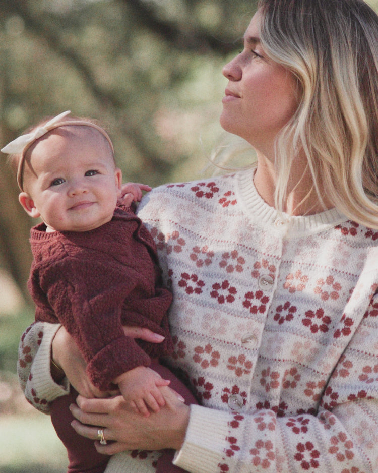 A woman holding a baby outdoors. The woman wears a cream knit cardigan with a red and pink floral pattern, and the baby wears a dark burgundy knit outfit and a white bow headband.