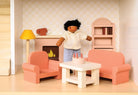 A miniature wooden dollhouse room interior featuring a wooden toy figure standing next to a coral-pink sofa and armchair set, a coffee table with a tiny red telephone, a pale pink decorative fireplace, a yellow floor lamp, and a natural wood bookcase against a patterned wallpaper background.