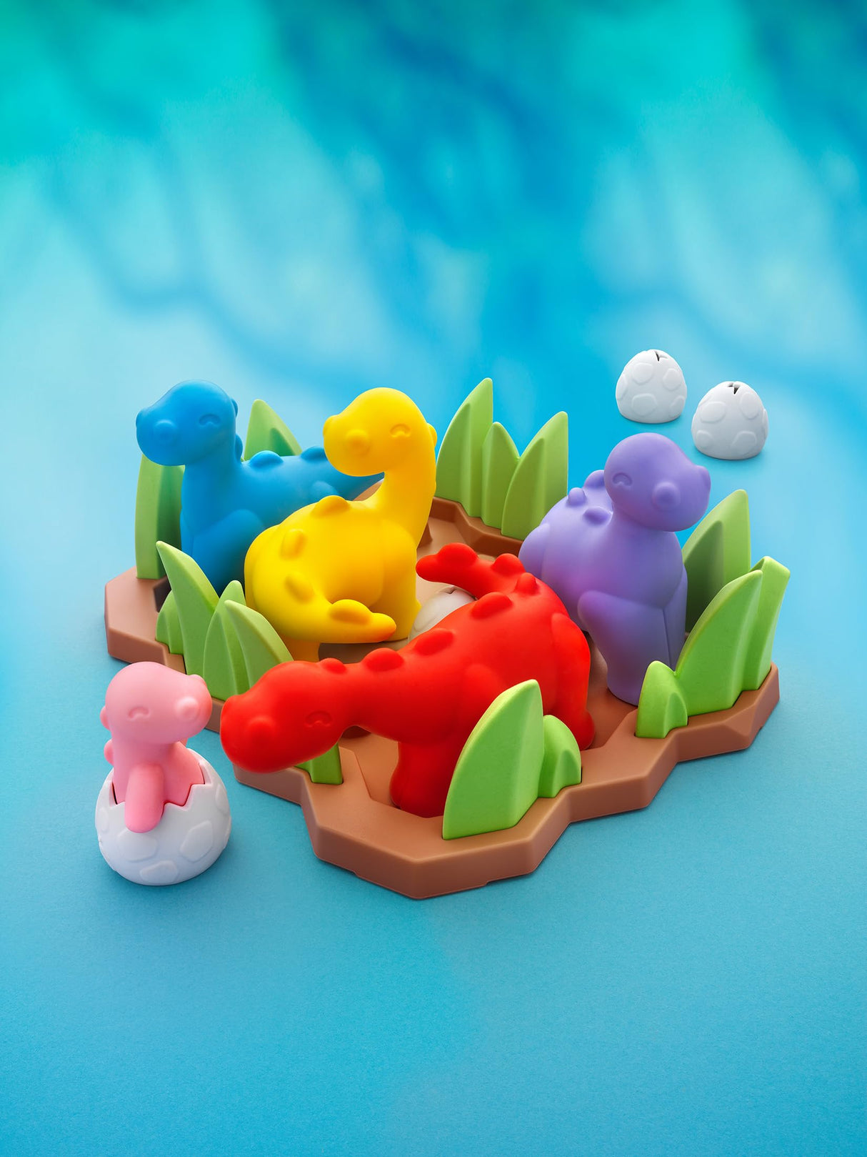 A colorful children's toy set featuring dinosaur figures and eggs on a brown base with green grass, photographed on a bright blue studio background.