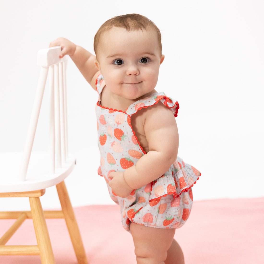 Baby in a heart-patterned outfit standing next to a chair on a white background