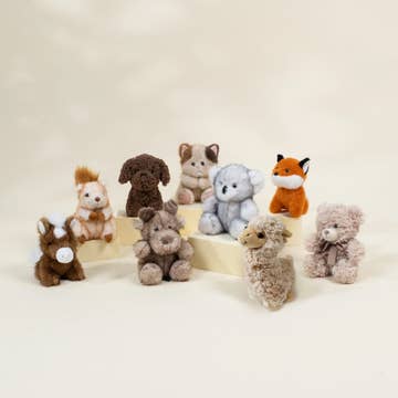 A group of small plush animal toys arranged on a light cream surface against a soft cream background. The toys include a brown horse, a white rabbit, a dark brown dog, a light brown cat, a light blue elephant, an orange fox, a gray sheep, and a beige bear.