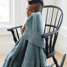 Person sitting on a black chair with a green blanket draped over them, in a bright room.