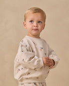 A toddler with blonde hair wearing a cream-colored long-sleeve sweatshirt with a small space-themed print of stars, planets, and rockets, posed against a neutral beige background.