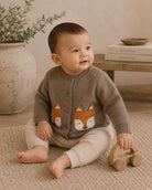A baby sitting on a textured rug wearing a brown button-up cardigan with a fox face design on the front, paired with cream pants. The baby is holding a wooden toy car in a cozy room setting.