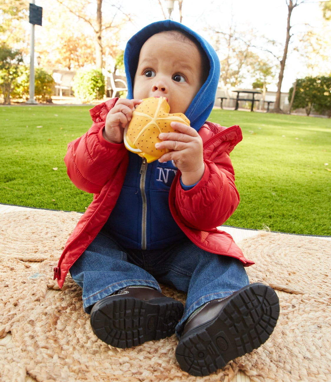 A baby sitting outdoors on a sandy surface wearing a blue and red outfit and holding a yellow and orange patterned toy.