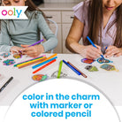 Two children sitting at a white table coloring in colorful charm stickers with markers and colored pencils. The Oooly logo is visible in the top left, and text at the bottom reads 'color in the charm with marker or colored pencil'.