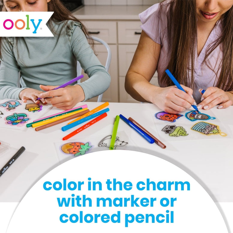 Two children sitting at a white table coloring in colorful charm stickers with markers and colored pencils. The Oooly logo is visible in the top left, and text at the bottom reads 'color in the charm with marker or colored pencil'.