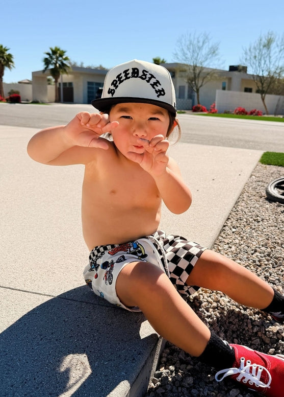 A toddler sitting outdoors on a concrete curb in front of a house, wearing a white and black checkered cap with 'SPEEDSTER' printed on it, white shorts with a black checkered pattern, black socks, and red sneakers.