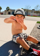 A toddler sitting outdoors on a concrete curb in front of a house, wearing a white and black checkered cap with 'SPEEDSTER' printed on it, white shorts with a black checkered pattern, black socks, and red sneakers.