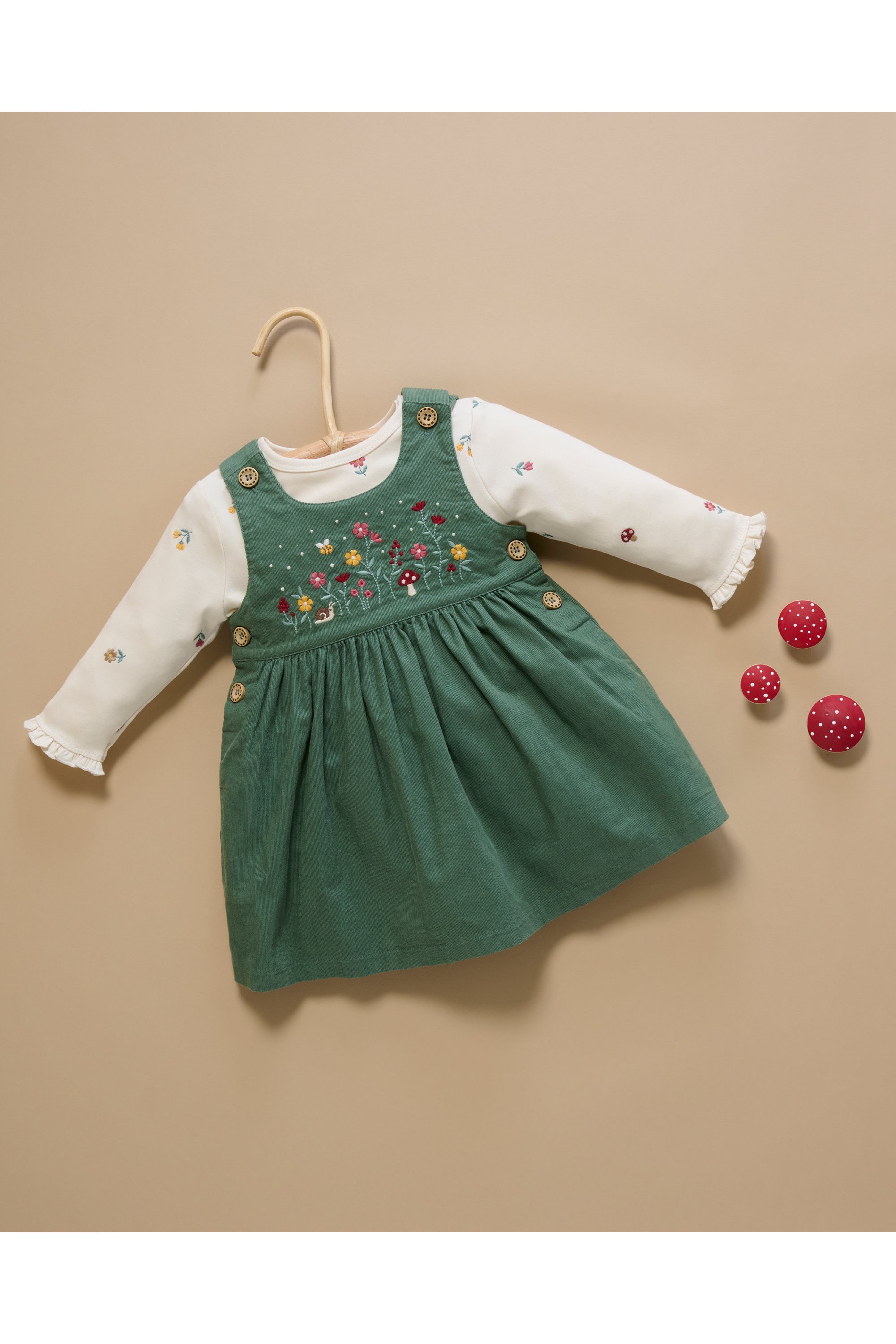 A toddler girl's long-sleeve dress featuring a sage green corduroy pinafore bodice with colorful floral embroidery and gold buttons, layered over a white long-sleeve top with ruffled cuffs. The dress is displayed on a wooden hanger against a solid beige background, with three small red pom-pom balls as styling props.