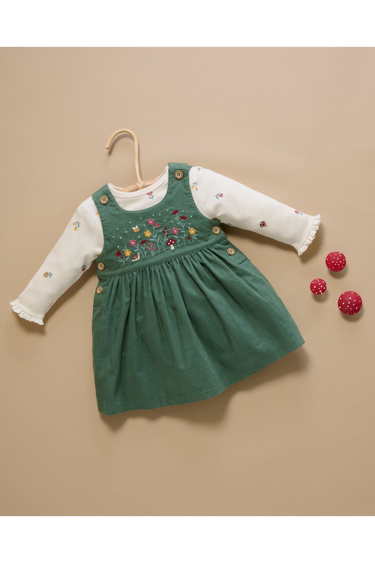 A toddler girl's long-sleeve dress featuring a sage green corduroy pinafore bodice with colorful floral embroidery and gold buttons, layered over a white long-sleeve top with ruffled cuffs. The dress is displayed on a wooden hanger against a solid beige background, with three small red pom-pom balls as styling props.