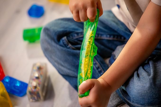 A child squeezes a long, translucent green Jelly Blox sensory building piece filled with small yellow beads, while other colorful blocks sit nearby on the floor.