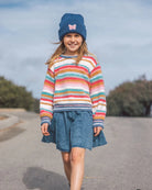 A young girl walking outdoors on a paved path wearing a colorful striped fuzzy knit sweater, a blue denim skirt, and a blue beanie with a pink bow.