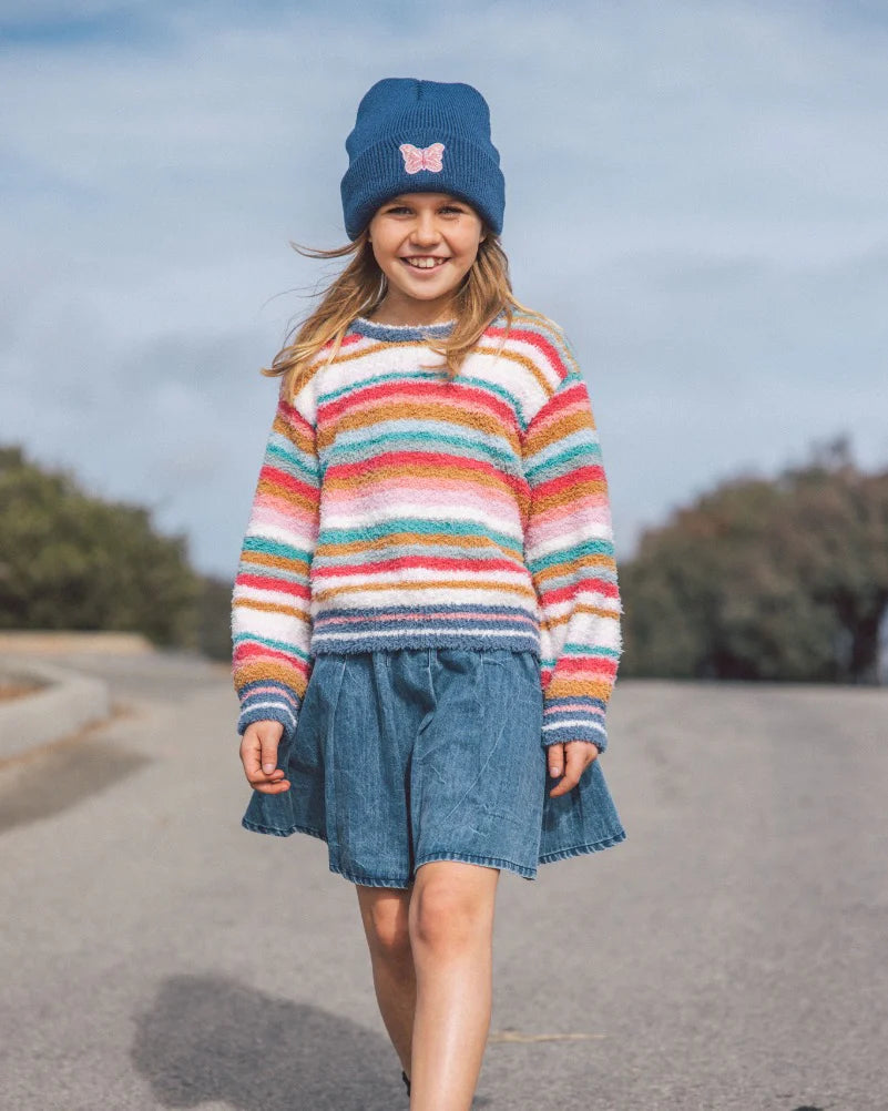 A young girl walking outdoors on a paved path wearing a colorful striped fuzzy knit sweater, a blue denim skirt, and a blue beanie with a pink bow.