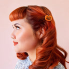 A woman in profile with bright red hair wearing a small orange pumpkin-shaped hair clip against a soft pink background.