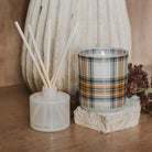 A reed diffuser bottle with white reeds and a lit glass candle in a plaid pattern, styled on a wooden surface with dried flowers and a textured cream vase in the background.