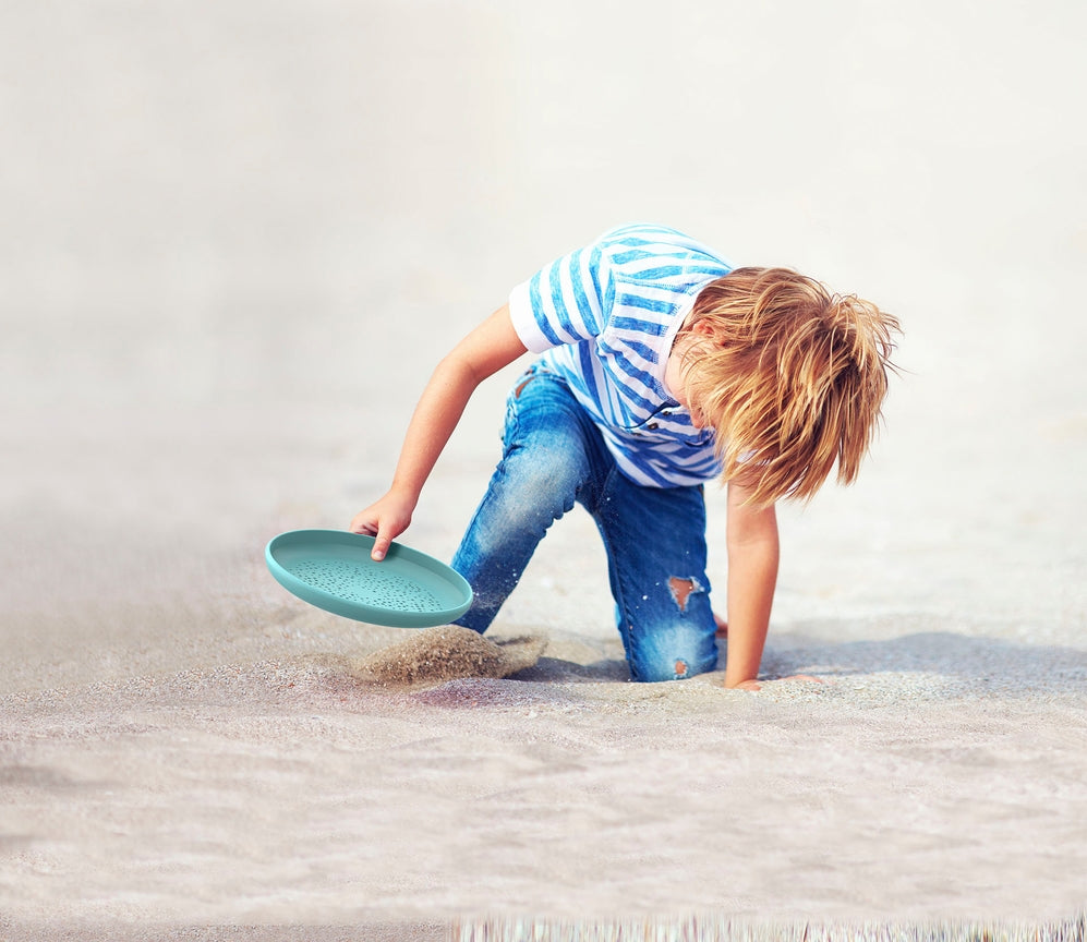 A child scooping sand into a turquoise flying disc on a beach.