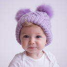 Baby wearing a purple knit hat with two pom-poms on a white background