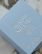 A close-up of a light blue textured perfume box with gold foil lettering that reads 'BOTANICA WINTER SOLSTICE SIGNATURE HOLIDAY ED.' on a light marble surface.