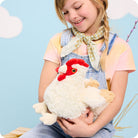 A young girl wearing overalls and a checkered scarf hugging a fluffy white Warmies chicken plush toy with a red comb and wattle.