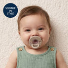 Baby wearing a green sleeveless dress with buttons, holding a beige pacifier, on a light textured background.