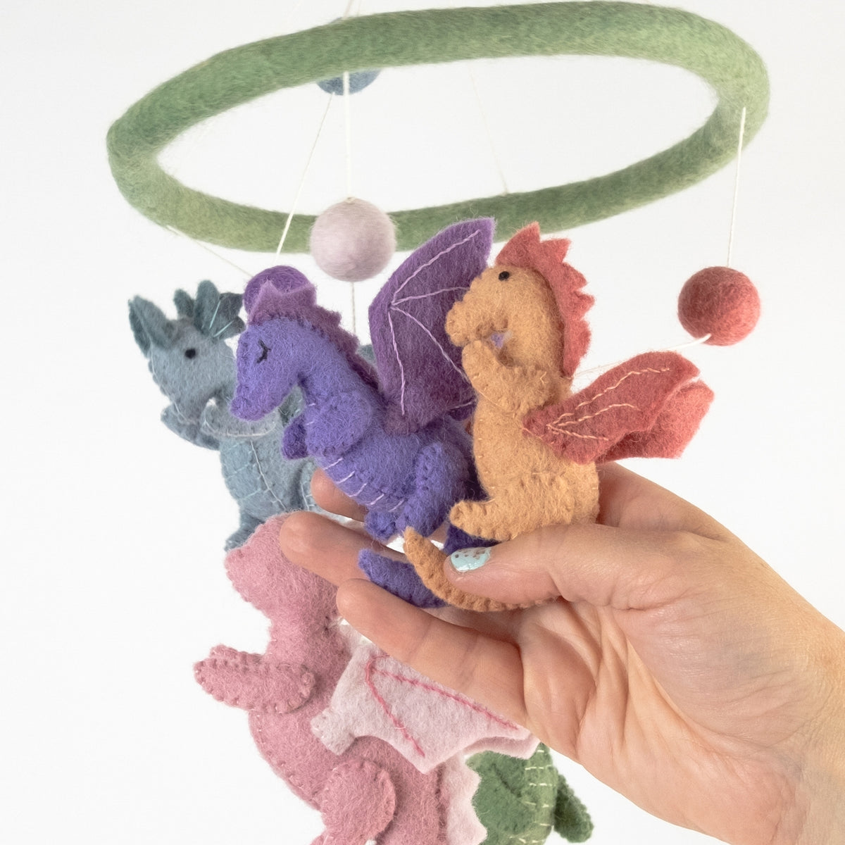 A hand holding a felt baby mobile with multiple colorful dragon figures hanging from a green circular ring against a white background.