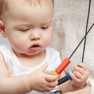 A baby wearing a white sleeve shirt holds a black silicone teether with a blue and tan accent against their cheek.
