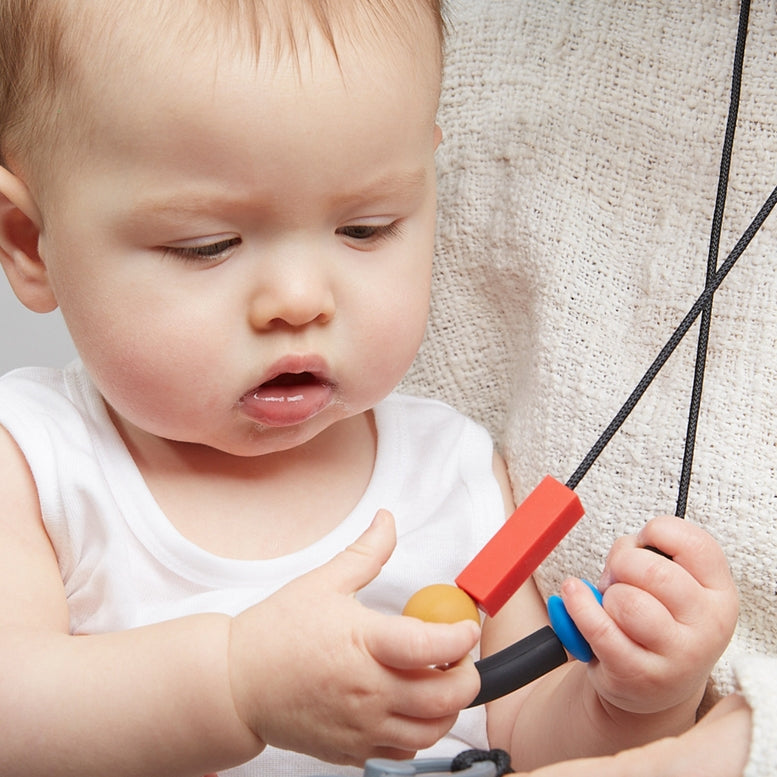 A baby wearing a white sleeve shirt holds a black silicone teether with a blue and tan accent against their cheek.