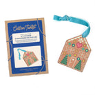 A gingerbread house shaped ornament shown next to its blue and kraft paper packaging card. The ornament features embroidered details in pink, green, yellow, and white on a tan background, with a light blue ribbon loop at the top.