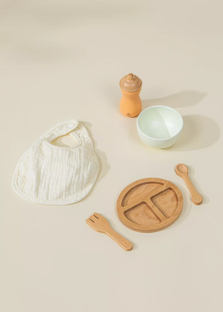 A baby feeding set arranged on a warm beige background, including a white muslin bib, a light wood divided plate, wooden utensils, a small white bowl, and an orange-toned bottle or dispenser.