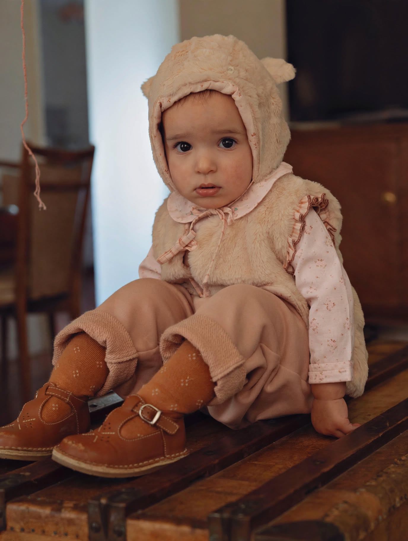 A baby sitting on a wooden bench wearing a beige teddy bear hooded outfit with a pink patterned long-sleeve shirt, beige pants, and brown leather shoes.