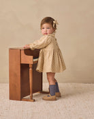 A toddler girl wearing a beige and white patterned long-sleeve dress, blue socks, and tan shoes stands next to a small wooden desk against a warm beige background.