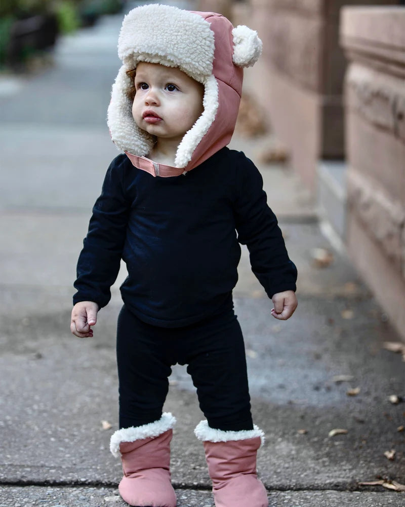 A toddler standing outdoors wearing a black outfit, pink winter boots with sherpa cuffs, and a pink and white sherpa-lined hat with bear ears.