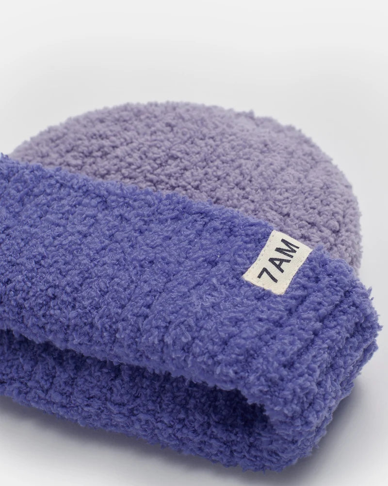 A two-tone ribbed fuzzy beanie in shades of purple (taro), featuring a folded brim with a small white tag branded with "7AM".