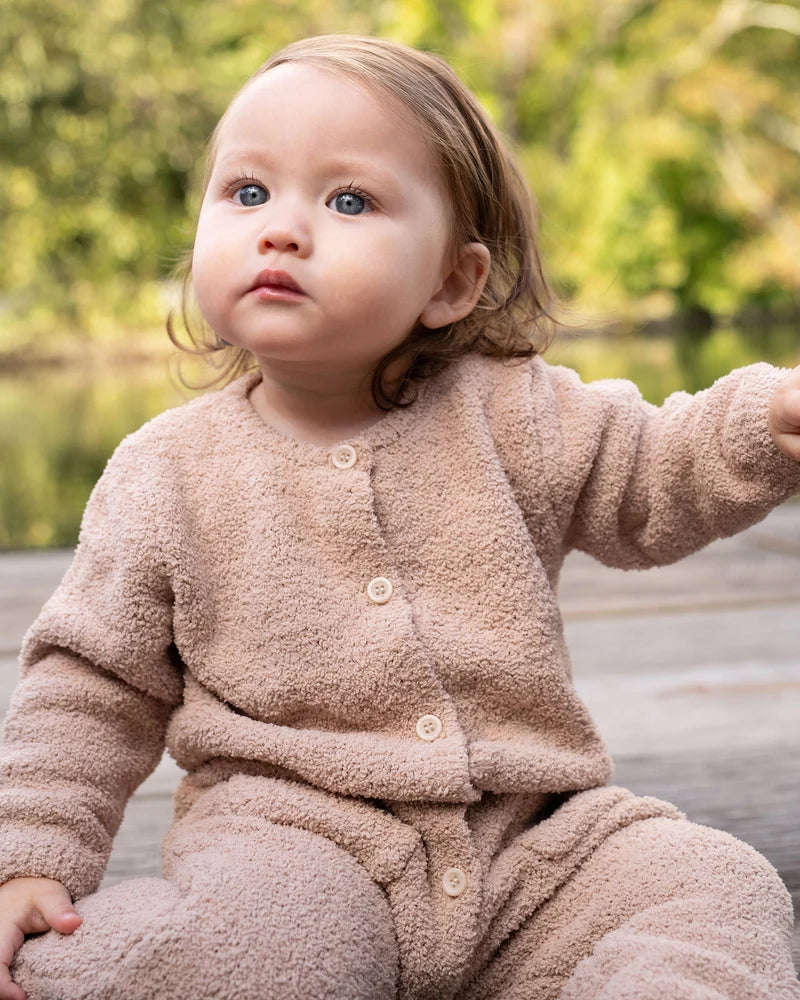 A toddler with reddish-brown hair sits outdoors in a natural, wooded setting, wearing a long-sleeve fuzzy romper in a warm pecan brown color by 7AM Enfant.