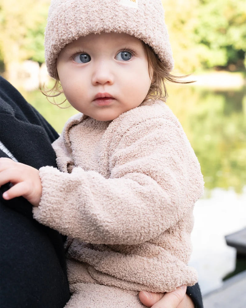A toddler wearing a matching fuzzy cap and a long-sleeve pecan brown romper by 7AM Enfant is being held outdoors.