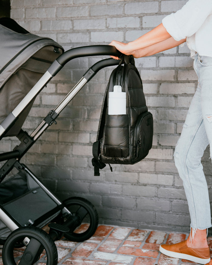 Person holding a black backpack next to a stroller against a brick wall.